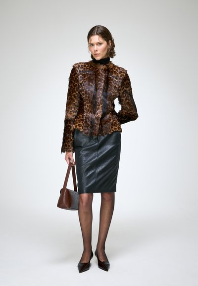 Femme portant une veste en fausse fourrure imprimé léopard, une jupe crayon en cuir noir, des talons noirs, et tenant un sac à main marron, debout devant un fond uni.