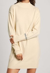 Abito maglione color crema con collo alto, polsini e orlo a costine. Presenta il logo "PEPE JEANS" sulla manica. Tessuto morbido.