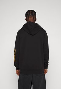 Sudadera negra con capucha grande. Las mangas largas tienen texto amarillo en el brazo izquierdo. Tejido suave, corte relajado y bolsillo tipo canguro.