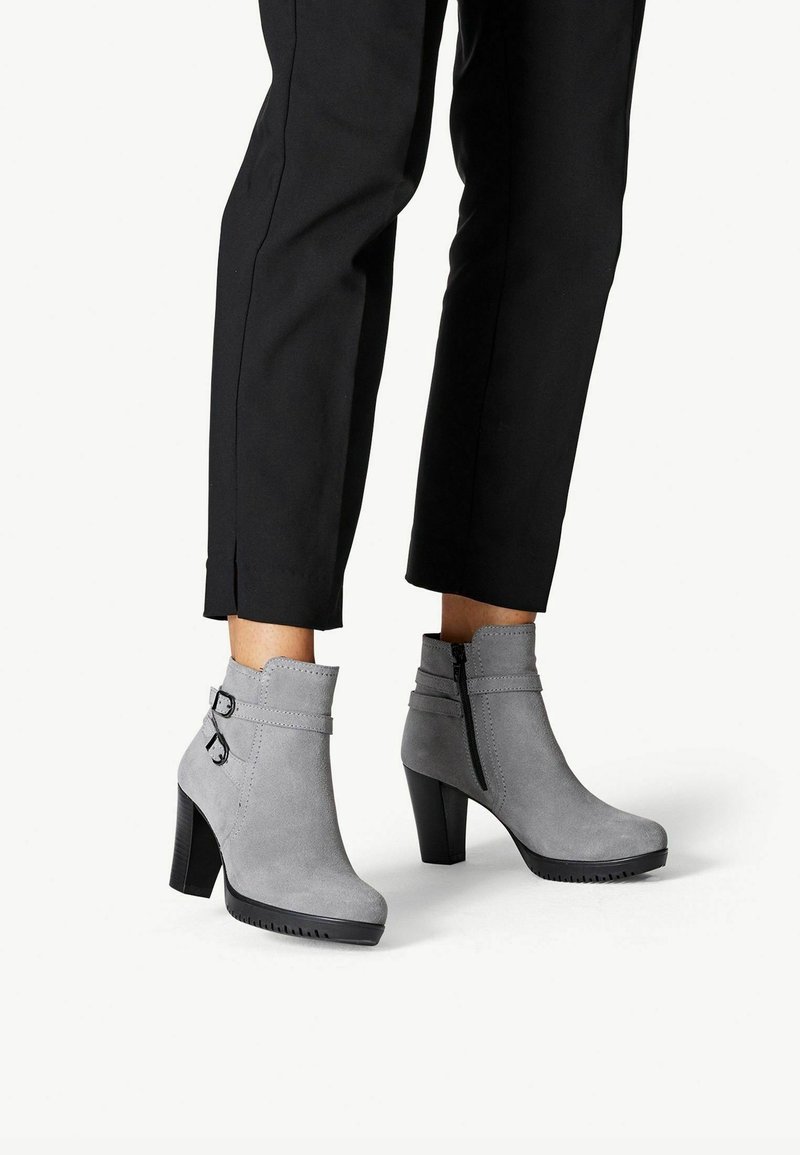 Personne portant un pantalon noir et des bottines gris suede avec talons carrés, fermeture éclair latérale et boucles décoratives.