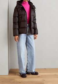 Doudoune noire à capuche, avec fermeture éclair et design matelassé. Portée par-dessus un col roulé rose vif et un jean à jambes larges bleu clair.