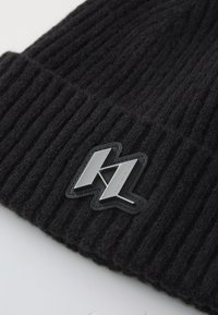 Căciulă neagră din tricot cu textură reliefată. Prezintă o etichetă logo argintie în față, evidențiind un design geometric și detalii de branding.
