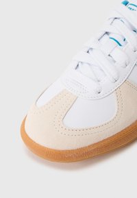 Białe sneakersy z gładkiej skórzanej cholewki, z zamszowymi akcentami w jasnym beżu i gumową podeszwą. Kryty sznurowany design z przeszyciami.
