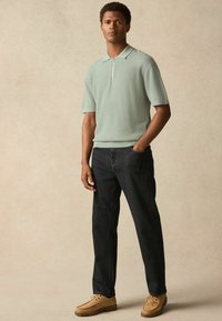 Mint green textured polo with a zip collar paired with dark jeans and beige shoes, standing against a neutral background.