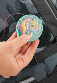 Gestickter Aufnäher mit einem Einhorn-Design und Regenbogenmähne, befestigt an einem strukturierten schwarzen Rucksack, der von einer Hand gehalten wird.