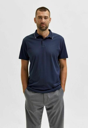 Camisa de polo navy con cuello que presenta acentos blancos, de mangas cortas y un corte ajustado, acompañada de pantalones grises.