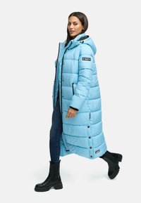 Lichtblauwe pufferjas met een capuchon, voorzien van brede horizontale stiknaden, zwarte accenten en drukknoopsluitingen, gecombineerd met zwarte chunky boots.
