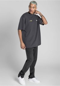 Př Oversized tmavě šedé tričko s kulatým výstřihem, s malým zlatým textem, spárované s černými džíny a bílými teniskami. Hladká textilie.