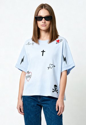 Femme aux cheveux raides portant des lunettes de soleil foncées, un T-shirt oversize bleu clair avec des motifs graphiques noirs et rouges, et un jean bleu.
