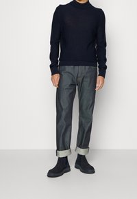 Marinblå turtleneck-tröja med ribbade cuffar och fåll, kombinerad med uppvikta mörka jeans och svarta anklarboots med en texturerad finish.