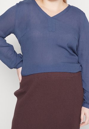 Femme portant un chemisier bleu à manches longues et une jupe marron côtelée, une main sur la hanche, sur un fond clair uni.