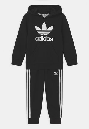 Felpa nera Adidas con grande logo bianco e tasca frontale, abbinata a pantaloni della tuta neri con strisce laterali bianche e logo.