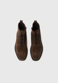 Botas de tobillo de ante marrón con punta redonda, detalle decorativo de brogue y cierre de cordones negro. Incluyen una cremallera lateral y suela texturizada.