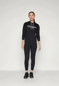 Frau steht da und trägt einen schwarzen Champion-Hoodie mit weißen Kordeln, schwarze Leggings und schwarze Sneaker vor einem schlichten hellen Hintergrund.