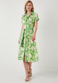 Grünes Blumenkleid mit kurzen Ärmeln, Knopfverschluss und Taillengürtel. Es hat ein Muster aus weißen Blumen auf einem lebhaften grünen Hintergrund.