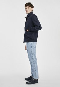 Granatowa kurtka zapinana na zamek z kołnierzem stojącym, elastycznymi mankietami i bocznymi kieszeniami, połączona z jasnoniebieskimi spodniami w prostym kroju i czarnymi sneakersami.