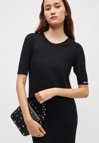 Μαύρο ριμπ φόρεμα με κοντά μανίκια, στρογγυλή λαιμόκοψη και λεπτομέρεια λογότυπου. Το μοντέλο κρατάει μια μαύρη τσάντα με τρουκς και ιμάντα ώμου.