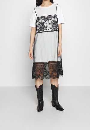 Femme portant une robe t-shirt blanche avec une superposition de robe nuisette en dentelle florale noire et des bottes western noires mi-mollet, debout sur un fond uni.