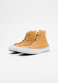 Wysokie trampki w musztardowym kolorze z płótna, z metalowymi oczkami. Biała gumowa podeszwa z fakturowanym wzorem i wzmocnionym noskiem.