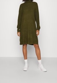 Person som bär en olivgrön långärmad stickad klänning, vita sneakers och vita strumpor, som står mot en enfärgad ljus bakgrund.