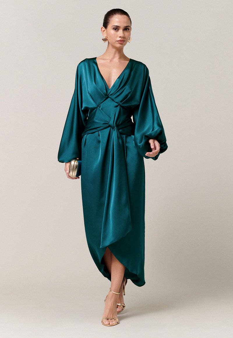 Femme portant une robe portefeuille en satin bleu sarcelle avec des manches longues bouffantes, des boucles d'oreilles anneaux dorés, tenant une pochette assortie et des sandales à talons hauts dorées.