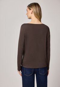 Bruine top met lange mouwen en een ronde hals, gemaakt van zachte stof. De achterkant heeft een rechte zoom en een losse pasvorm. Gecombineerd met een blauwe spijkerbroek.