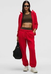 Červená mikina s kapucí a odpovídající tepláky z měkké látky, doplněné hnědým crop topem. Doplňky zahrnují černou tašku a sluneční brýle.