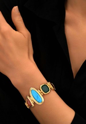 Bracelet en or avec de grandes pierres turquoise, vertes et brun rougeâtre porté au poignet sur un fond de vêtement noir.