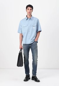 Lys blå kortermet skjorte med knapper og to frontlommer, kombinert med falmede jeans og svarte skinnsko, holder en svart bag.
