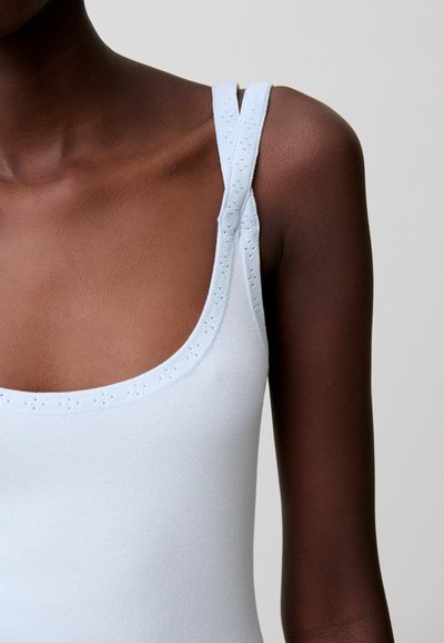Persona dalla pelle scura che indossa un top senza maniche azzurro pallido con spalline decorative traforate, mostrata dalla clavicola alla parte superiore del braccio su uno sfondo neutro.