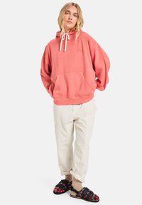 Oversized coral hoodie with drawstring, front pocket, casually styled with beige cargo pants and black plaid sandals.