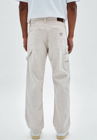 Hombre con pantalones cargo beige de pierna ancha con múltiples bolsillos y zapatillas blancas, de pie contra un fondo claro y liso.