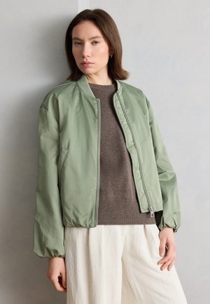 Γυναίκα με ίσια καστανά μαλλιά, φοράει ανοιχτό πράσινο μπουφάν τύπου «μπόμερ» πάνω από καφέ πουλόβερ και άσπρο παντελόνι, στέκεται μπροστά σε ανοιχτό γκρι τοίχο.