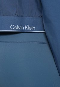 Modré športové oblečenie s hladkou, elastickou textúrou. Pás má biely logotyp "Calvin Klein" s tenkým bielym ozdobným lemom.
