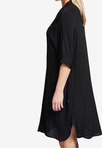 Svart, knälång klänning med lös passform, korta ärmar, sidslitsar och en mjuk textur. Har en subtil lyster och minimalistisk design.