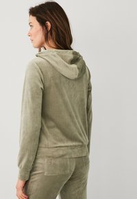 Haut à capuche en polaire de couleur olive douce, doté d'une capuche avec cordon, de manchettes ajustées et d'une texture lisse, associé à un pantalon assorti.