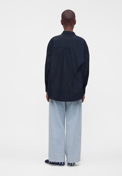 Personne portant une chemise ample sombre à manches longues et un jean clair à jambes larges avec des ourlets effilochés, debout de dos devant un fond clair uni.