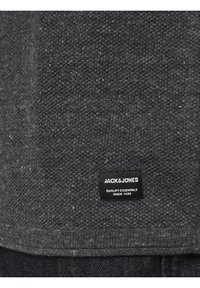 Ourlet de pull en maille gris foncé avec une étiquette noire portant la mention « JACK & JONES QUALITY ESSENTIALS SINCE 1990 » au-dessus d'un jean en denim foncé.