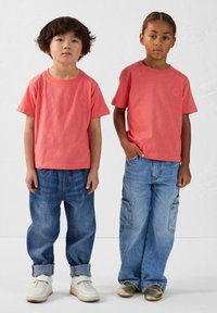 Deux enfants portent des t-shirts en coton corail assortis et des jeans en denim bleu ample. L'un porte des baskets blanches, l'autre des chaussures dorées.