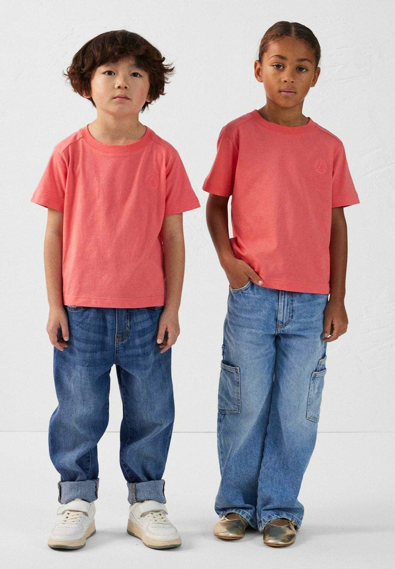 Deux enfants portent des t-shirts en coton corail assortis et des jeans en denim bleu ample. L'un porte des baskets blanches, l'autre des chaussures dorées.