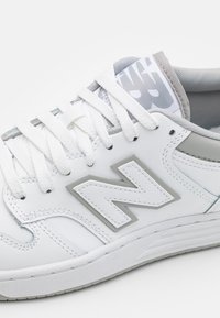 Białe skórzane sneakersy z szarymi akcentami, teksturowaną podeszwą i wyraźnym logo "N". Zawierają białe sznurówki i wzorzysty język.