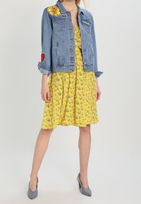 Veste en jean avec broderies florales et en forme de cœur, associée à une robe jaune à fleurs. La tenue est complétée par des talons hauts gris pour un look coordonné.