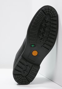 Suela de goma negra con surcos profundos, con un logo verde y naranja, parte superior de cuero liso y forma redondeada adecuada para uso casual.