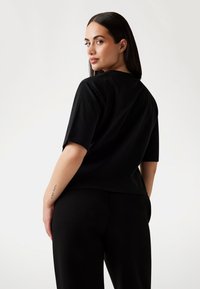 Žena s tmavými vlasy, oblečená v černém topu s krátkým rukávem a kalhotách, otočená mírně zpět, ukazuje tetování na zápěstí pravé ruky.