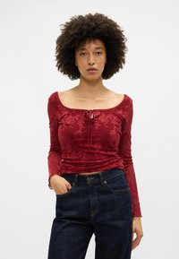 Κόκκινη μπλούζα μακρύ μανίκι με λουλουδένιο σχέδιο, διαθέτει στρογγυλή λαιμόκοψη και κορδόνι μπροστά. Συνδυασμένη με σκούρα μπλε τζιν ψηλόμεσο.