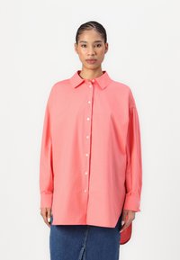 Chemise à manches longues rose corail avec col, présentant une coupe décontractée, des fentes latérales et des poignets retroussés. Fabriquée en tissu de coton léger.