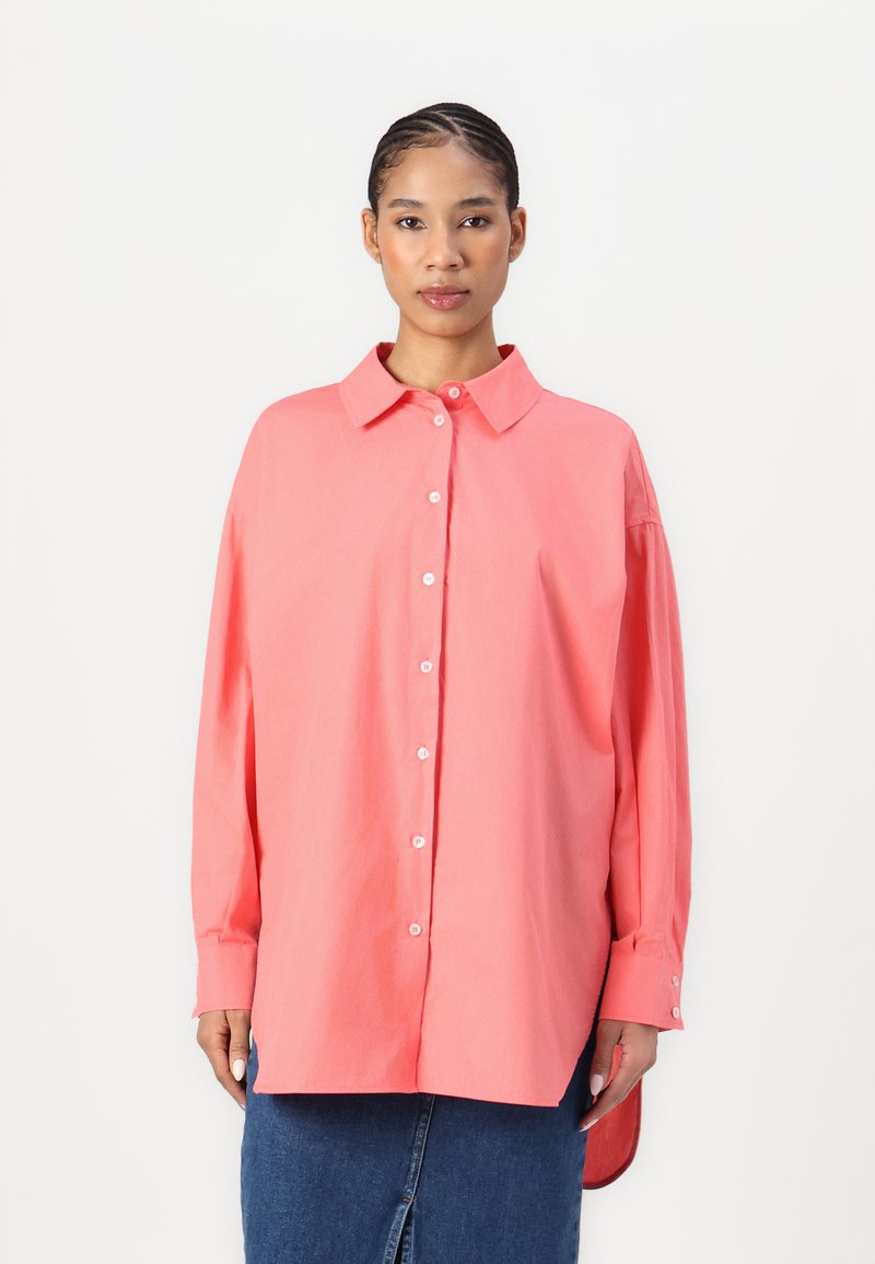 Camicia a maniche lunghe di colore rosa corallo con colletto, caratterizzata da una vestibilità rilassata, spacchi laterali e polsini arrotolati. Realizzata in tessuto di cotone leggero.