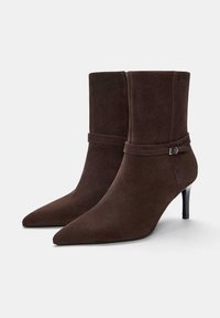 Stivaletti alla caviglia in suede marrone scuro con punta affilata, dotati di un tacco sottile e una cintura decorativa con dettaglio in fibbia argentata.