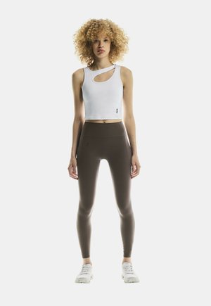 Mladý človek s kučeravými blond vlasmi, ktorý má na sebe biely crop top s výrezmi, tmavohnedé legíny a biele tenisky, stojí čelom.