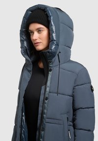 Polstret grå jakke med en stor, quiltet hætte, lynlås foran og flere lommer. Stoffet har en glat, skinnende tekstur. Justerbare snore.